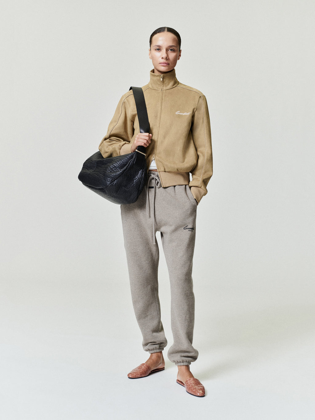 Person wearing a beige jacket and gray pants holding a black bag on a white background