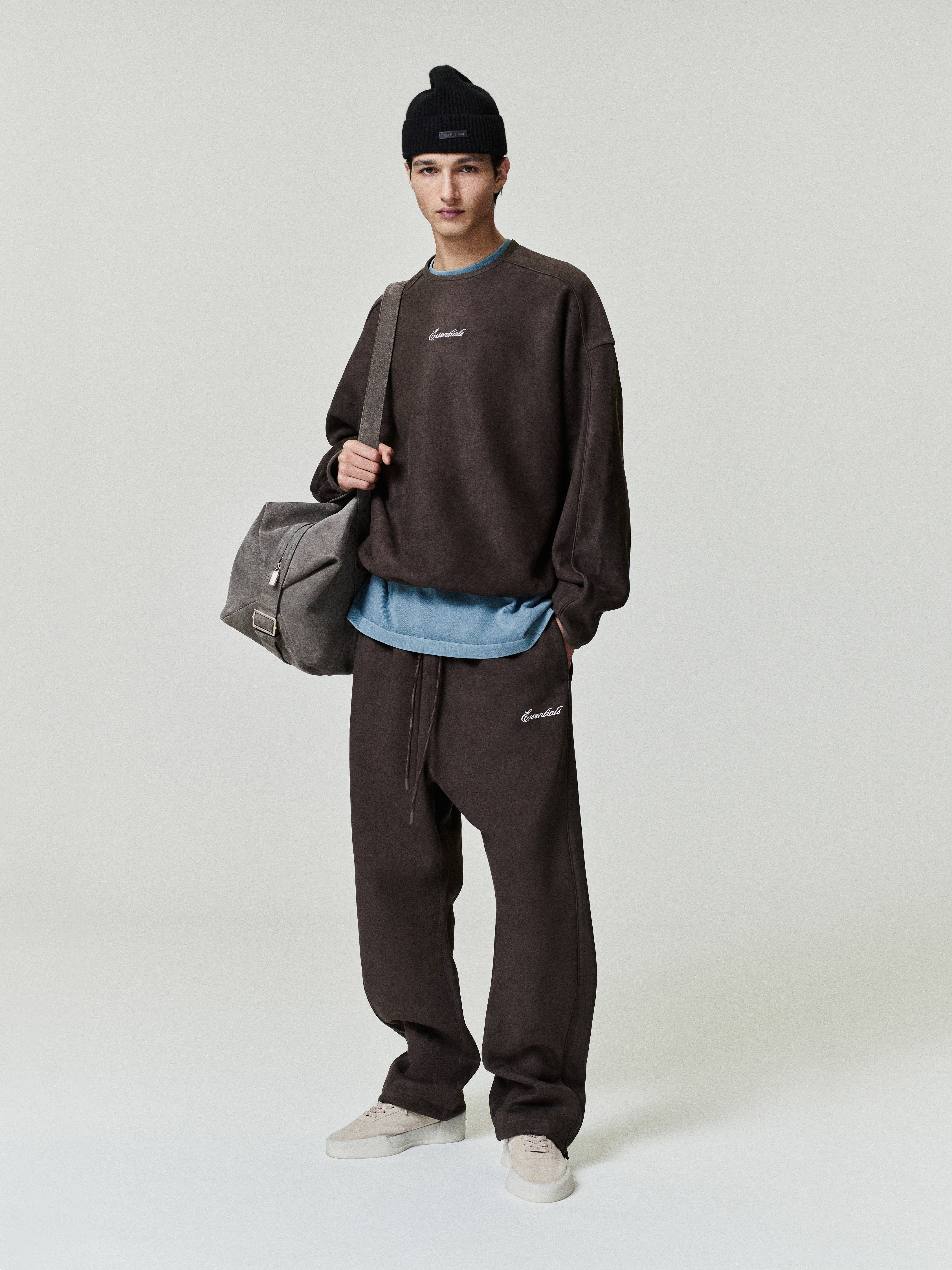 Person wearing a brown sweatshirt and pants with a gray bag on a white background