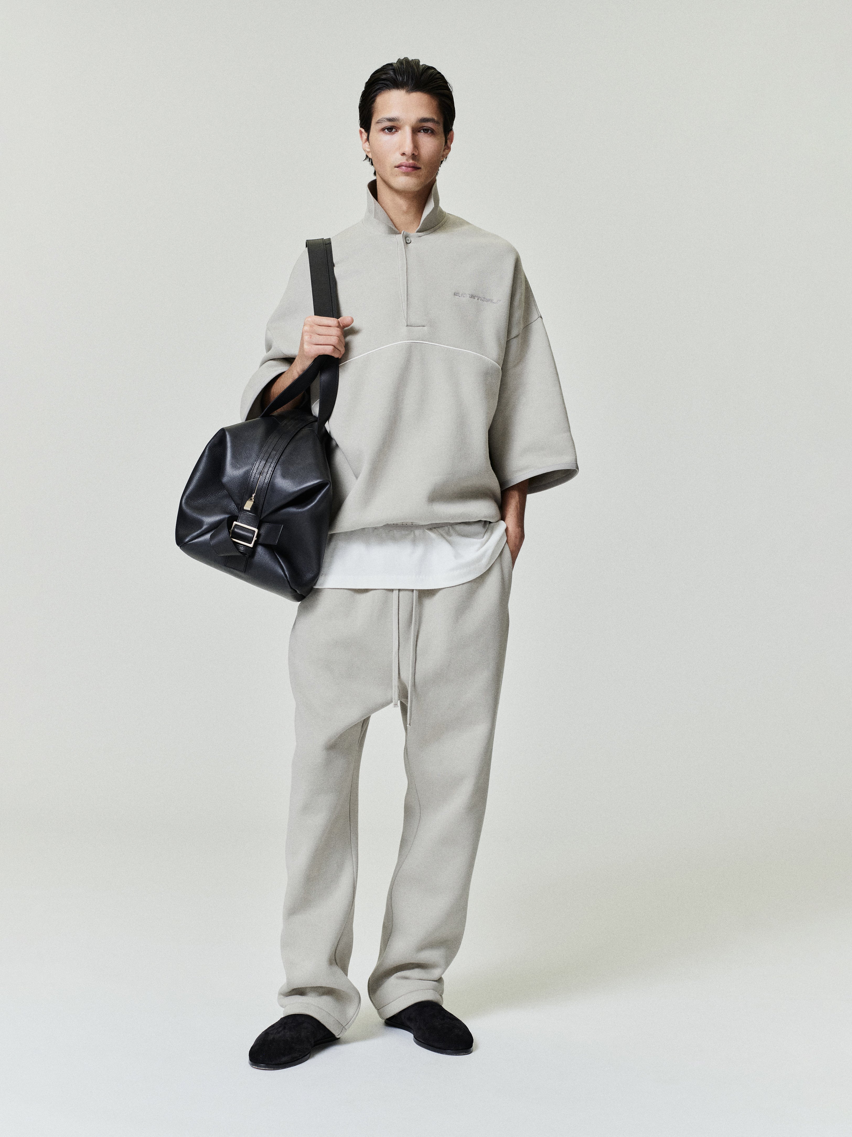 man wearing light gray cropped fleece polo sweatshirt with wide 3/4 sleeves and matching relaxed fleece pants carrying black leather bag