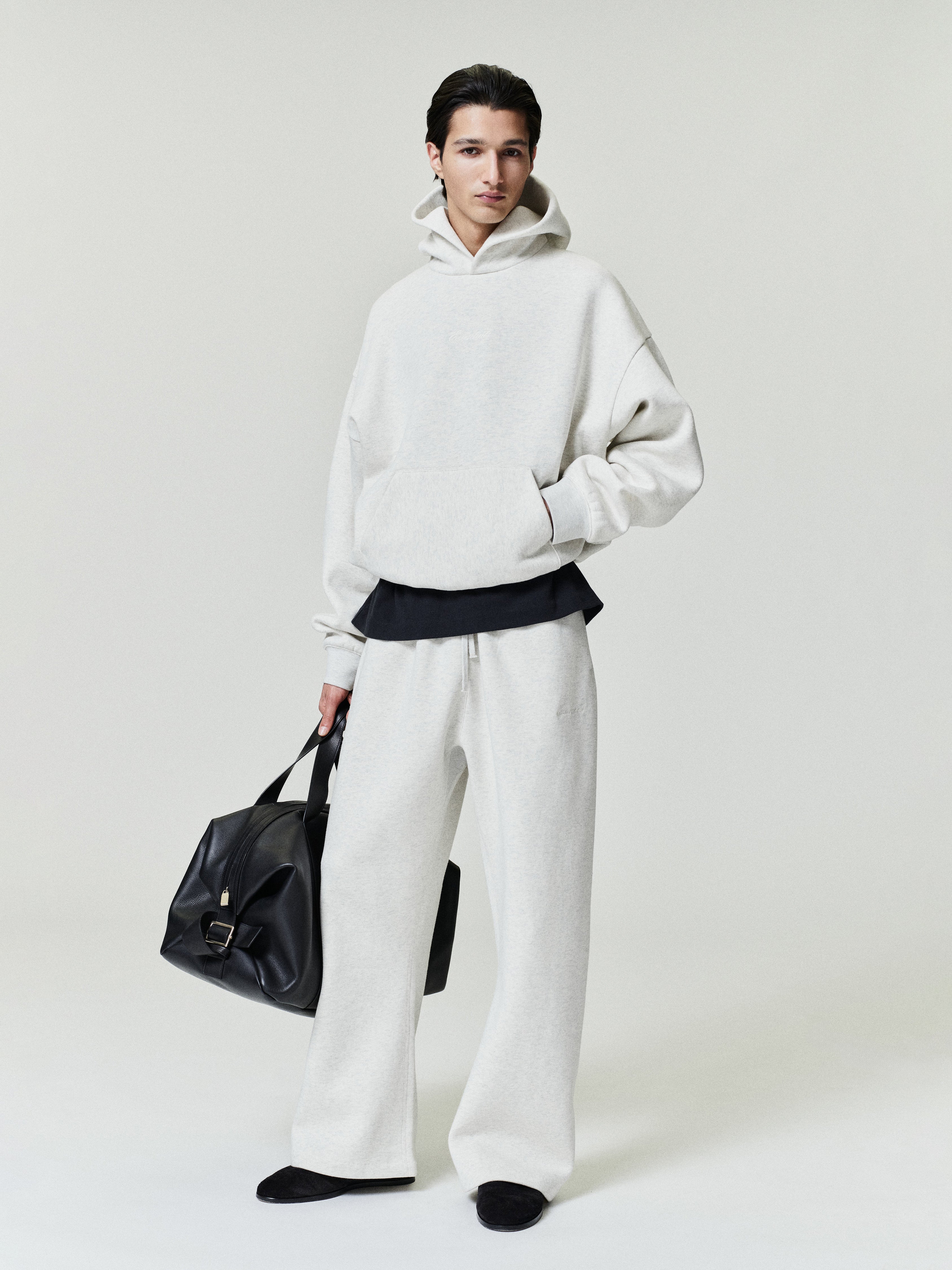 Person wearing a white hoodie and pants holding a black bag on a plain background